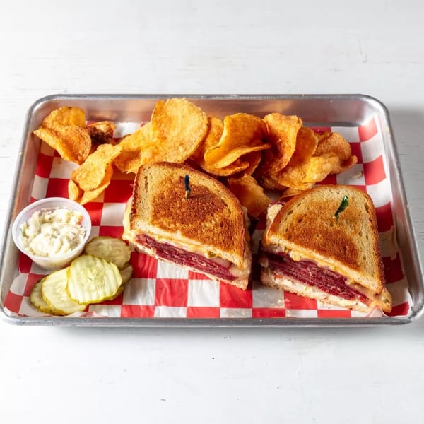 A reuben sandwich cut in half, with potato chips, coleslaw, and pickles on a red and white checkered tray.