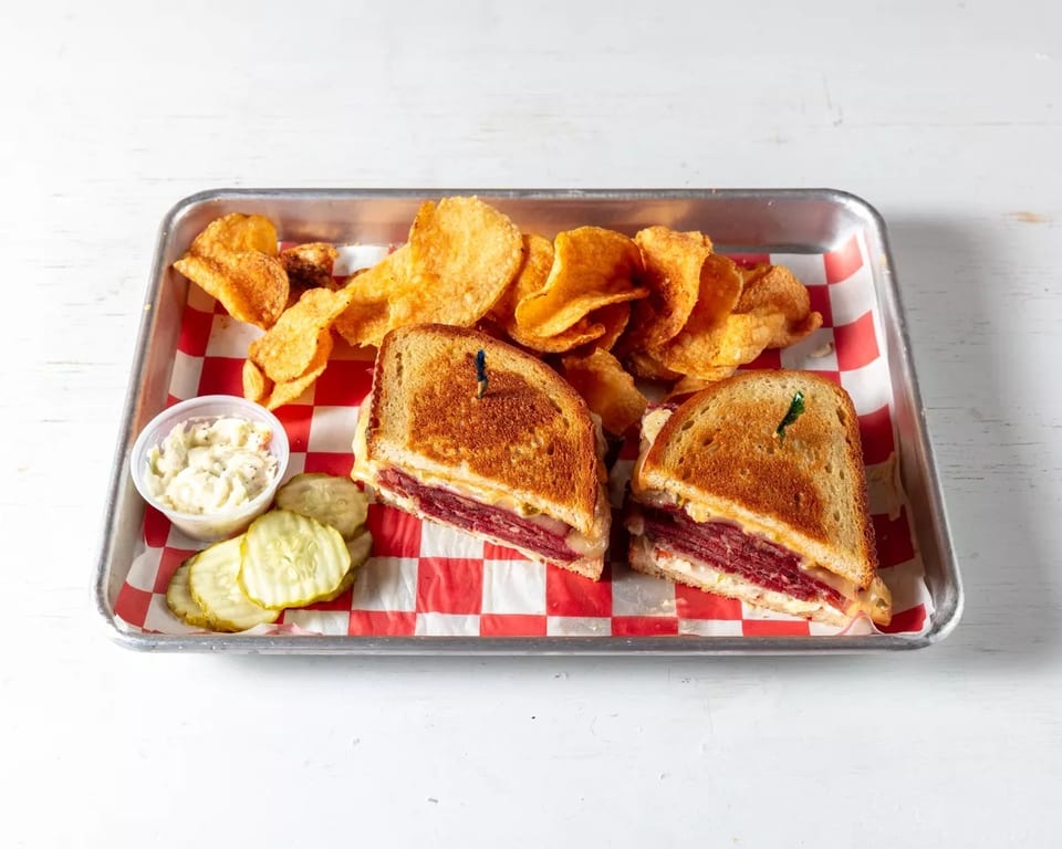 A reuben sandwich cut in half, with potato chips, coleslaw, and pickles on a red and white checkered tray.
