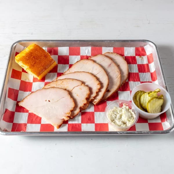 Sliced turkey, cornbread, coleslaw, and pickles on a red and white checkered tray.