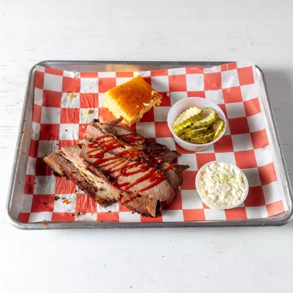 Smoked brisket with BBQ sauce, cornbread, pickles, and coleslaw on a checkered paper-lined tray.