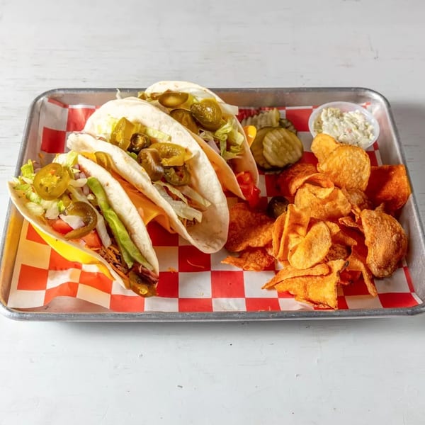 Three tacos with chips, pickles, and coleslaw on a metal tray with red and white checkered paper.