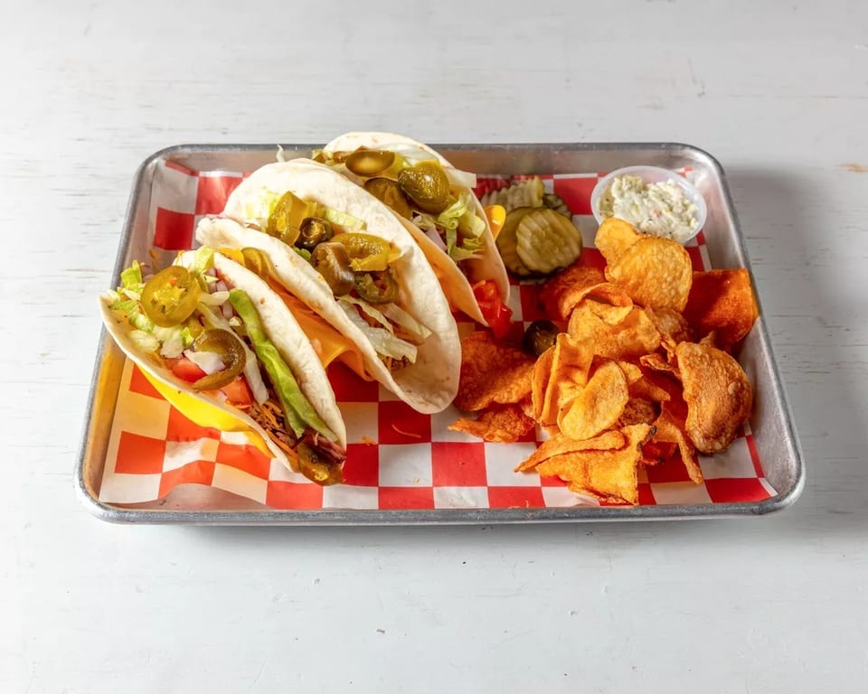 Three tacos with chips, pickles, and coleslaw on a metal tray with red and white checkered paper.