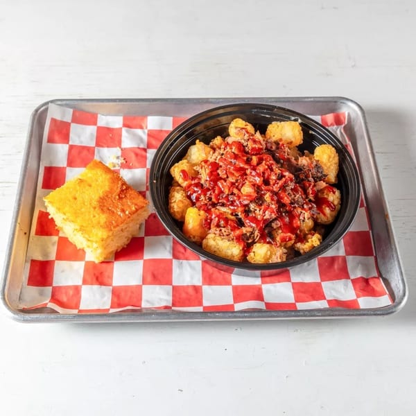 A tray with a bowl of tater tots topped with pulled pork and BBQ sauce, and a piece of cornbread.