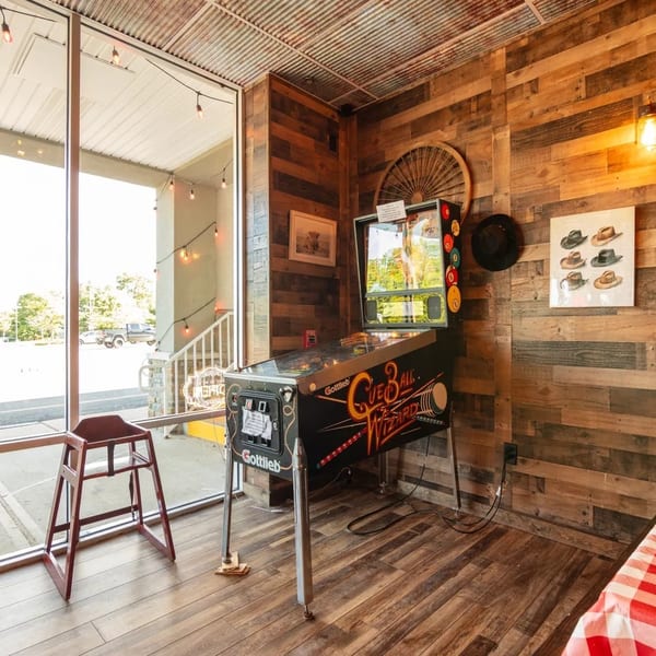 A vintage pinball machine stands next to a large window in a rustic room with wood-paneled walls.