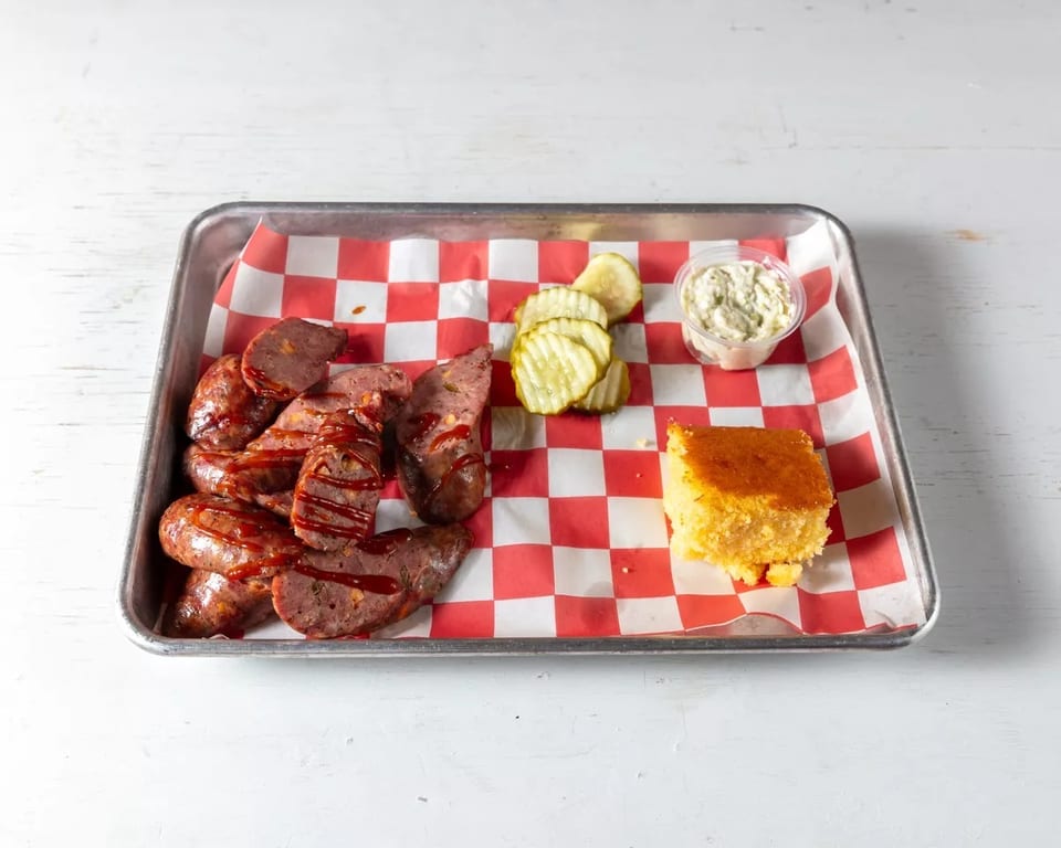 Sausage with barbecue sauce, pickles, cornbread, and a side of sauce on a checkered tray.