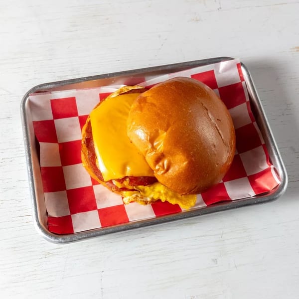 Breakfast sandwich with cheese, egg, and meat on a bun, served on a tray with checkered paper.