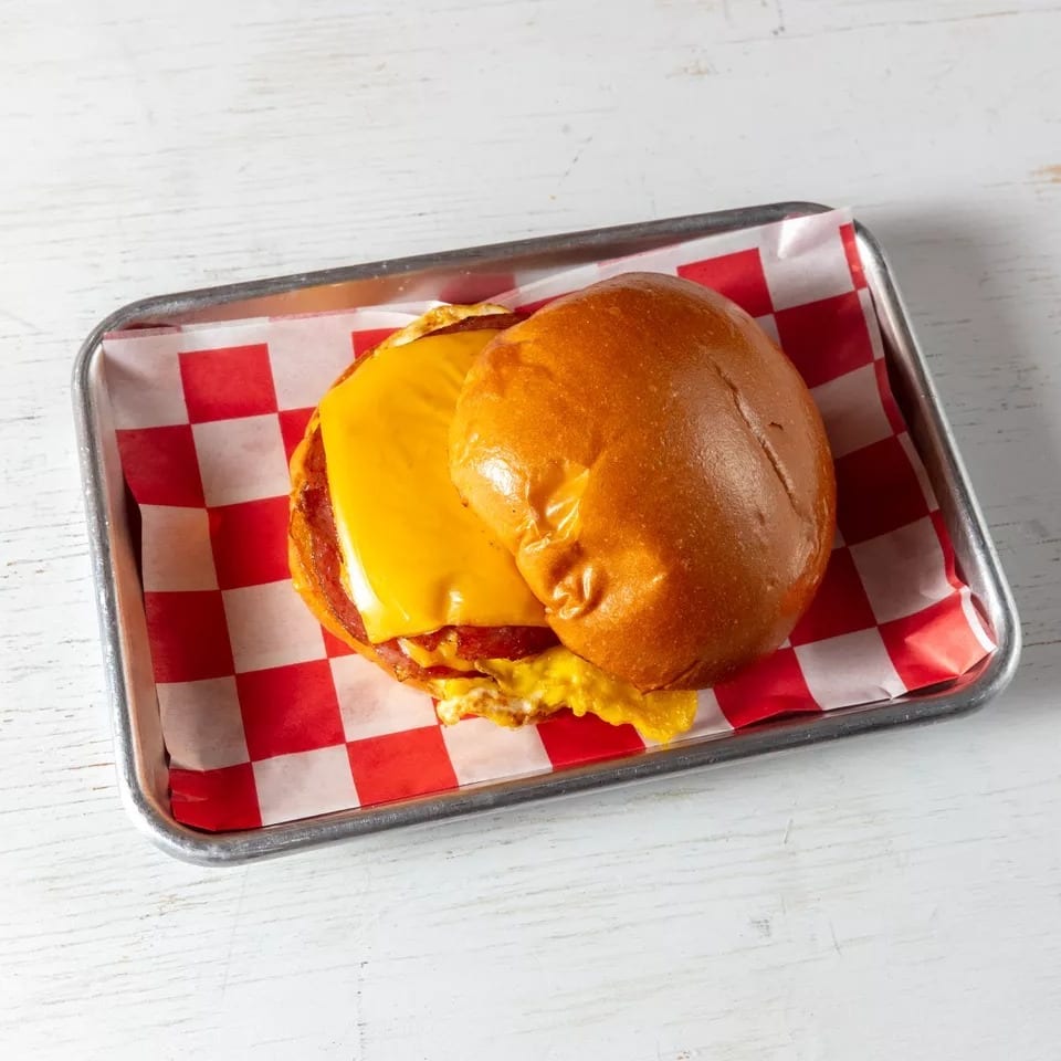 Breakfast sandwich with cheese, egg, and meat on a bun, served on a tray with checkered paper.